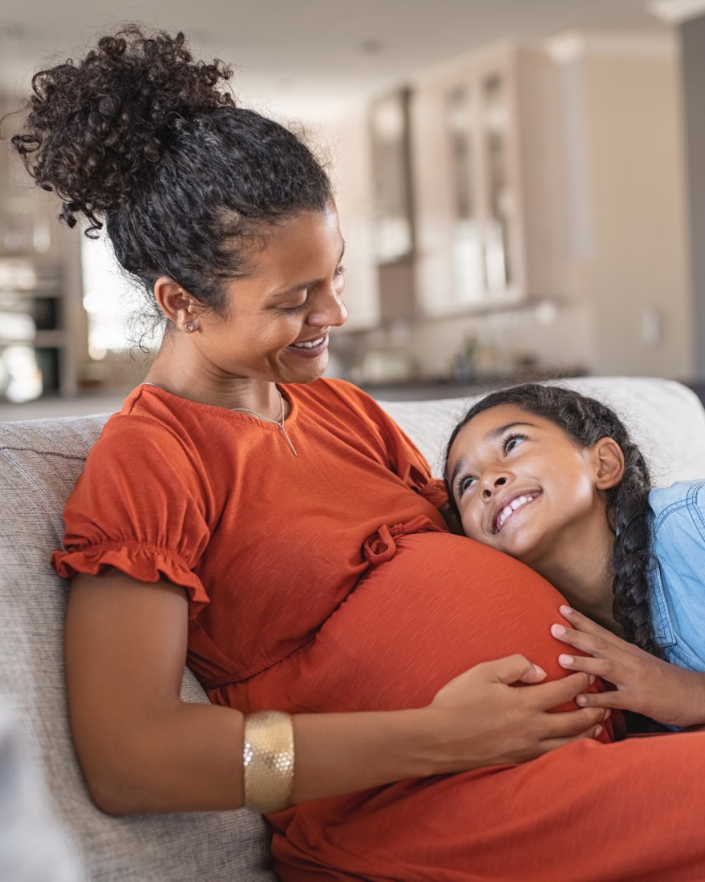 A pregnant woman on a sofa at home, smiling down at her daughter, who rests her head on her belly.