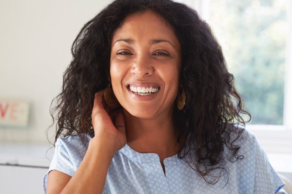 A woman in her forties seated in a bright room at home, laughing toward the camera.