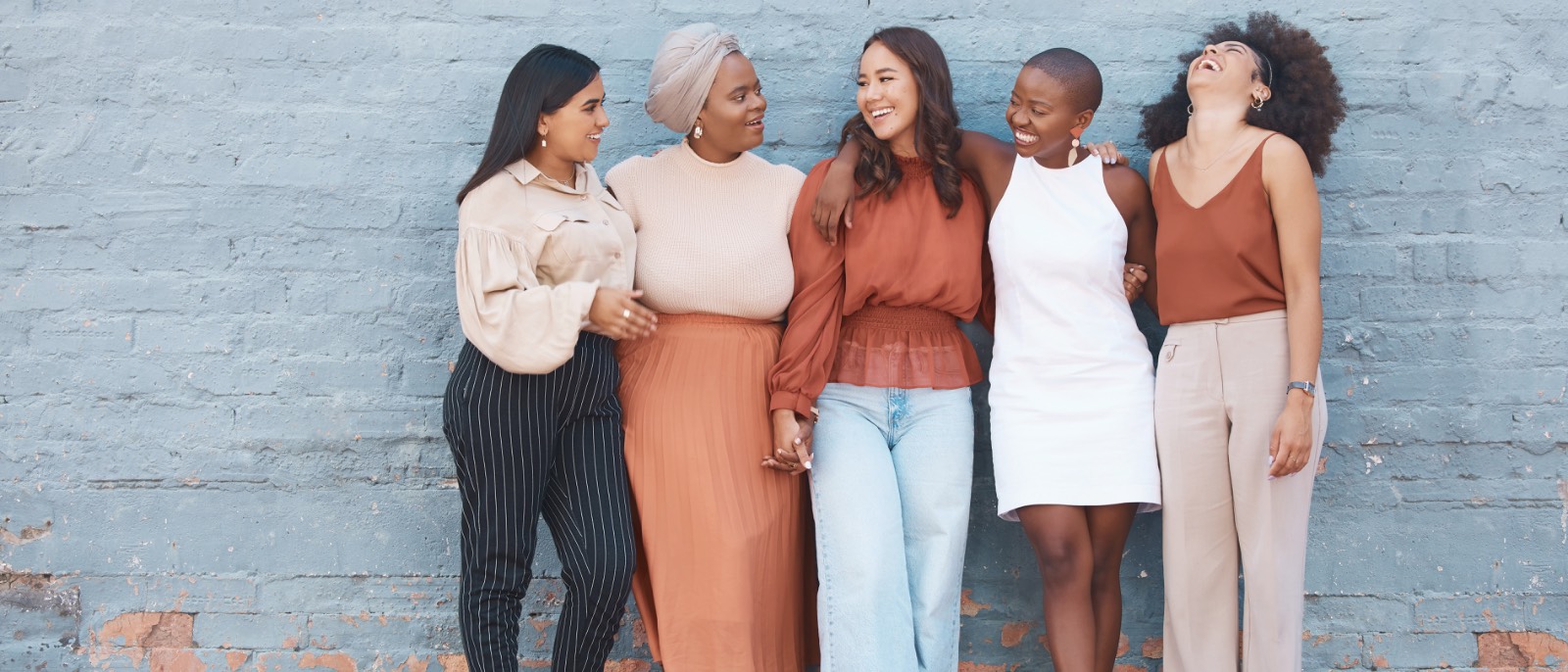 Five women standing together against a painted brick wall, arms linked, laughing.